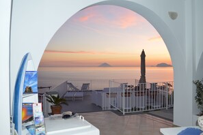 A terrace with a view of the sea at sunset, featuring a statue, lounge chairs, and a white railing. - A Alera Room and Breakfast (Malfa)