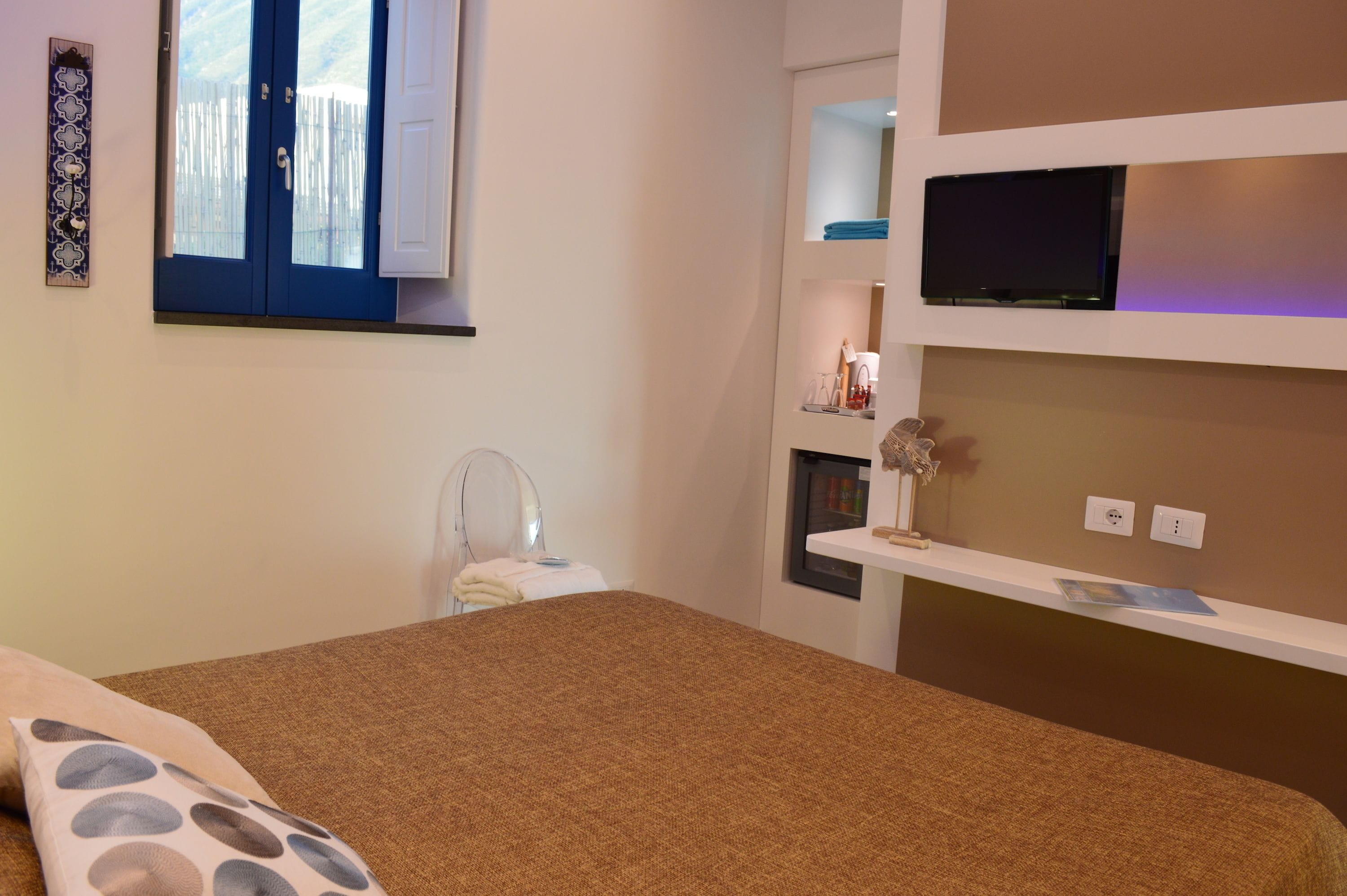 A bedroom with a bed, a TV mounted on the wall, a small table with a lamp, and a window with a blue door.