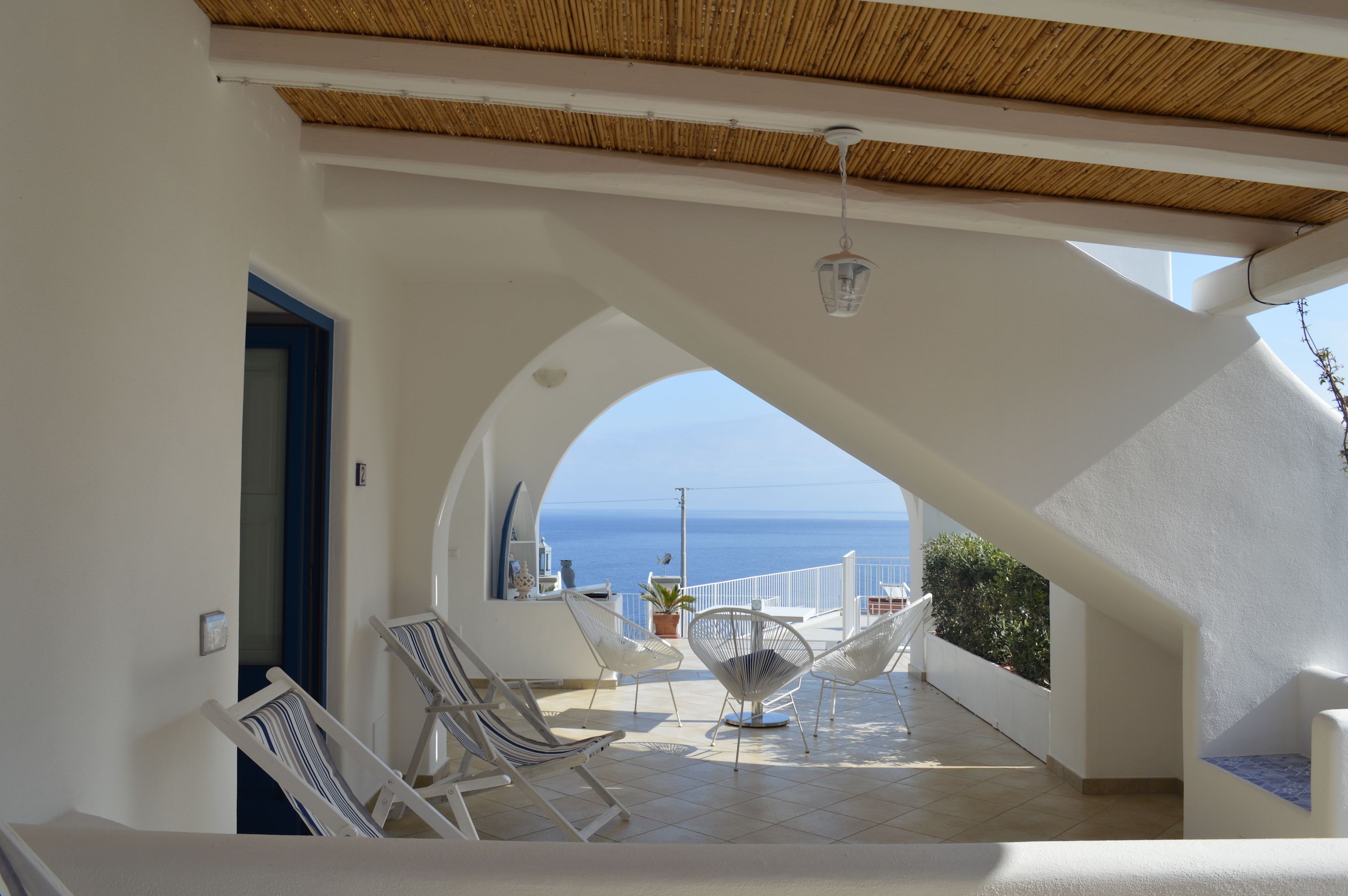 A covered outdoor seating area with a view of the sea, white walls, and a thatched roof.