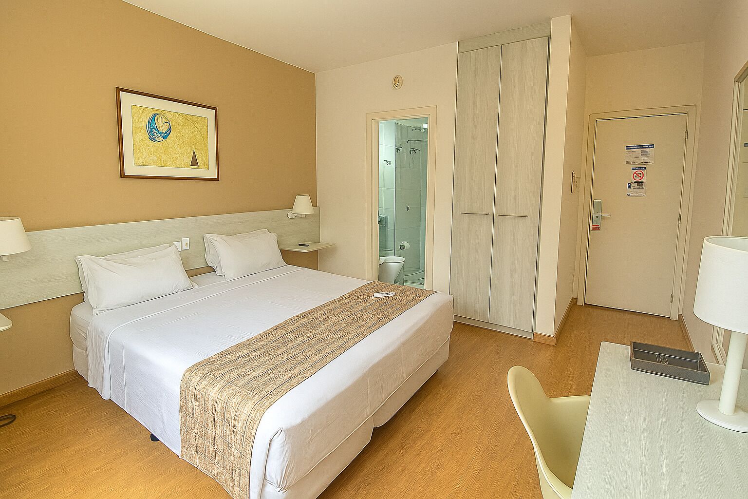 Une chambre d’hôtel comprenant un lit, des tables de chevet, un bureau, une chaise, une armoire et une salle de bain visible à travers une porte ouverte.