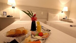 Habitación de hotel con una bandeja de desayuno ordenada sobre la cama, que incluye tostadas, bollería, una ensalada de frutas y un vaso de zumo de naranja.