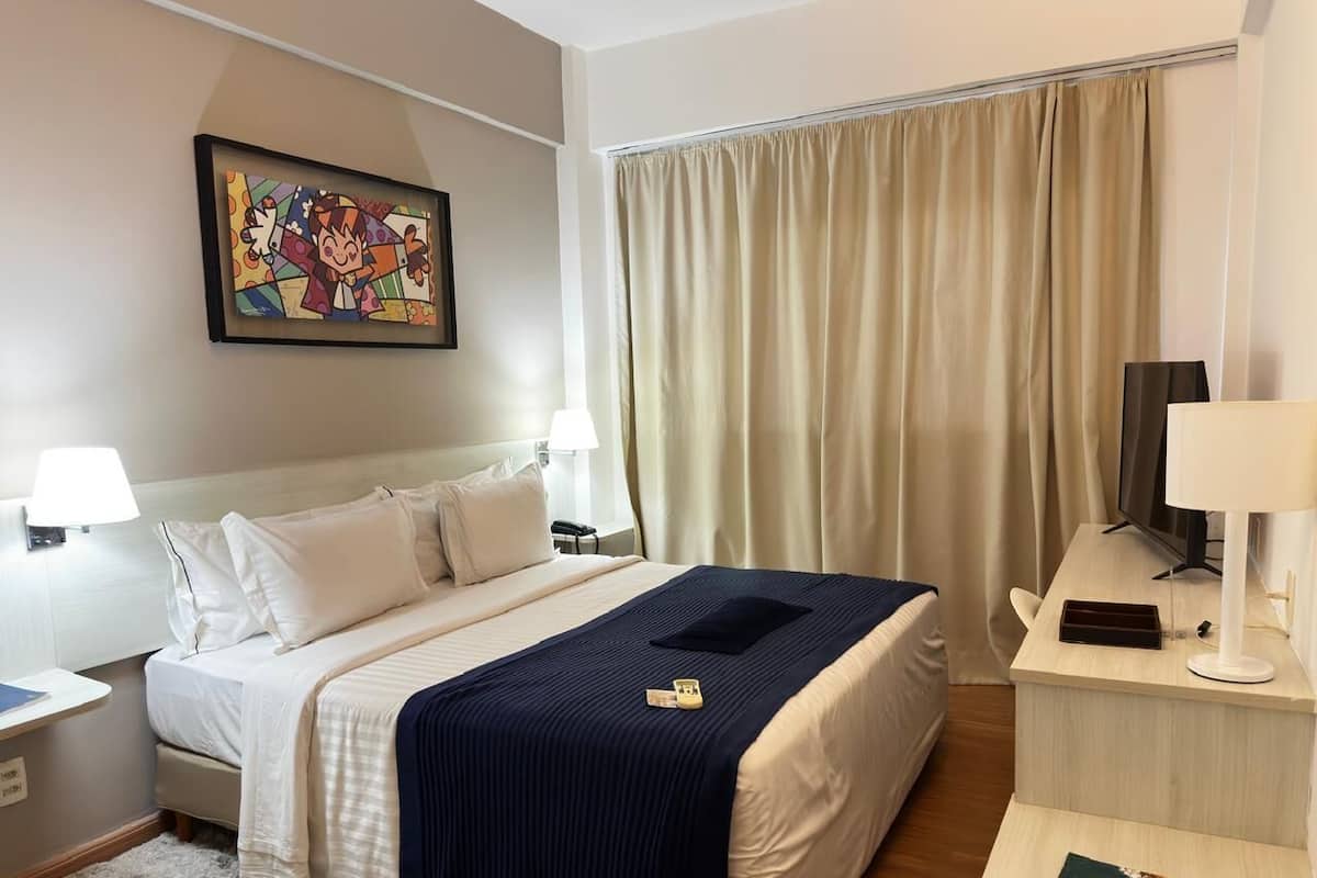 Habitación de hotel con una cama, dos lámparas de noche, un escritorio con lámpara, televisor y un cuadro en la pared.