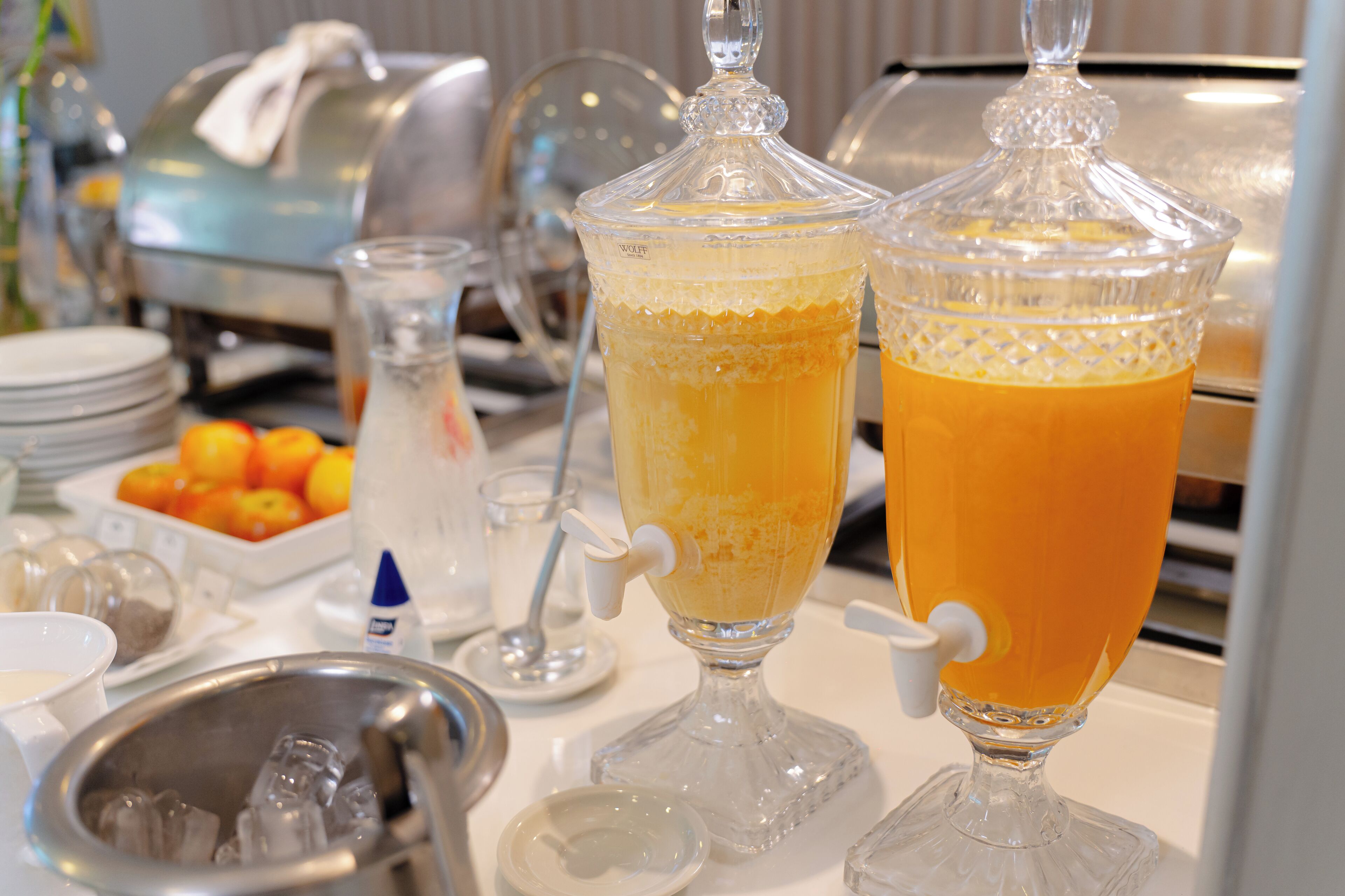 Un buffet comprenant deux grands récipients remplis de jus d’orange, un saladier de fruits et divers plats de service.