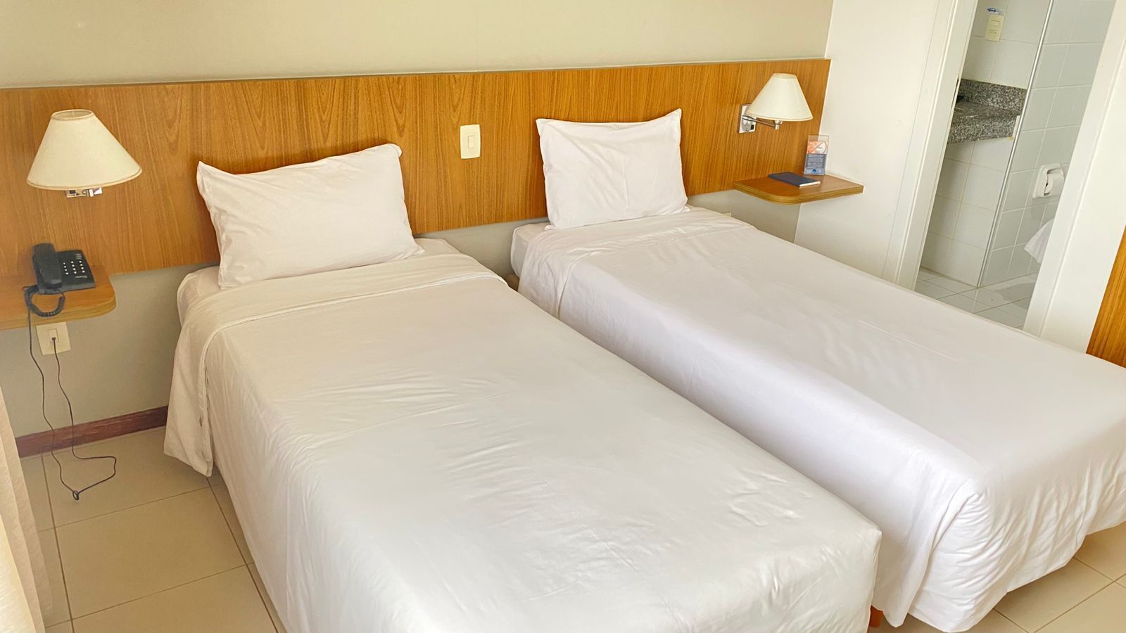 A hotel room with two single beds, a wooden headboard, bedside lamps, a phone, and a small table with a book and a lamp.