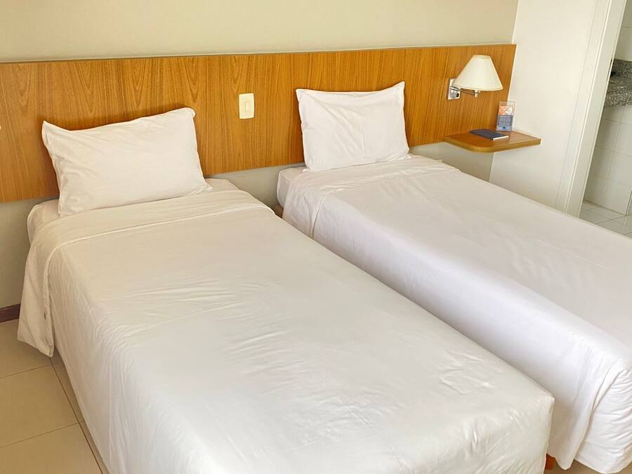 Habitación de hotel con dos camas individuales, cabecera de madera, lámparas de noche, un teléfono y una mesita con un libro y una lámpara.