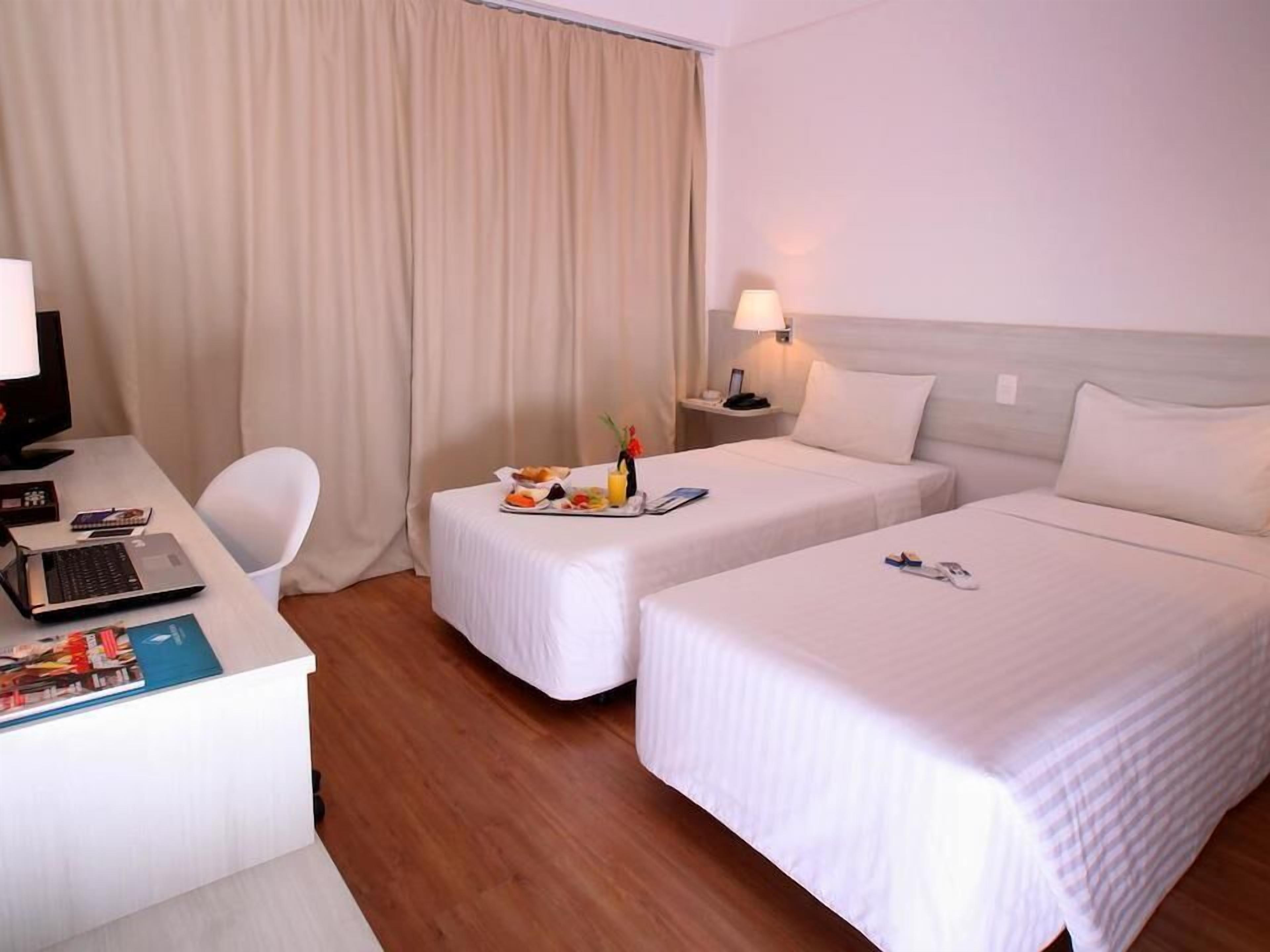 Ein Hotelzimmer mit zwei Betten, einem Schreibtisch mit Laptop, einem Stuhl und einem kleinen Tisch mit Frühstücksgebäck.