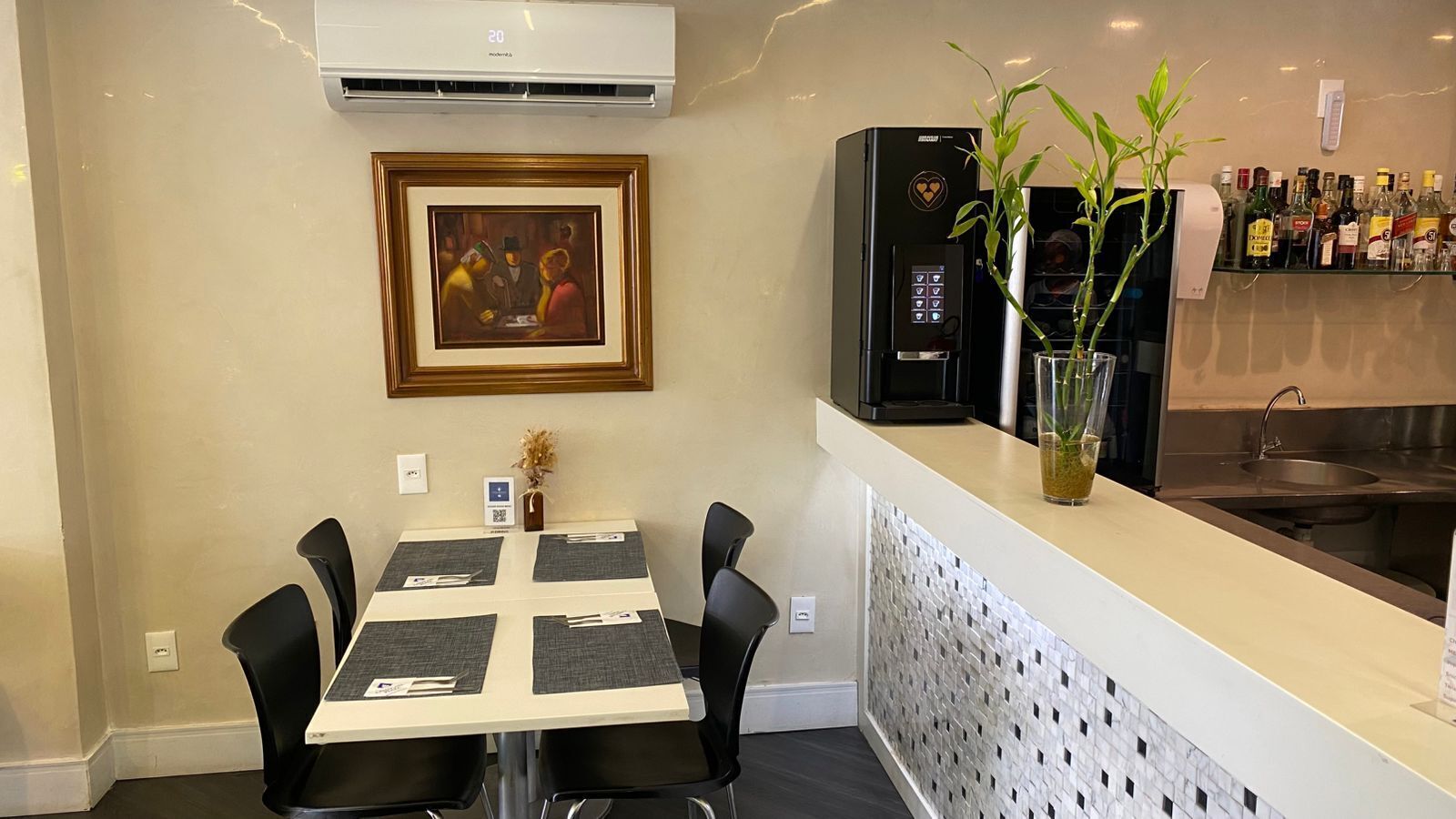 A dining area with a table set for four, a framed painting on the wall, a coffee machine, and a bar area with various bottles.