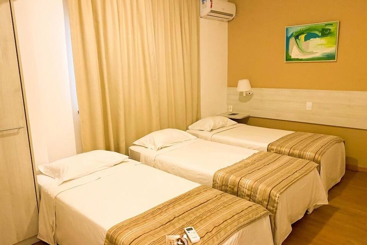 Habitación de hotel con dos camas individuales, un ventanal con cortinas, un equipo de aire acondicionado, una lámpara de noche y un cuadro en la pared.