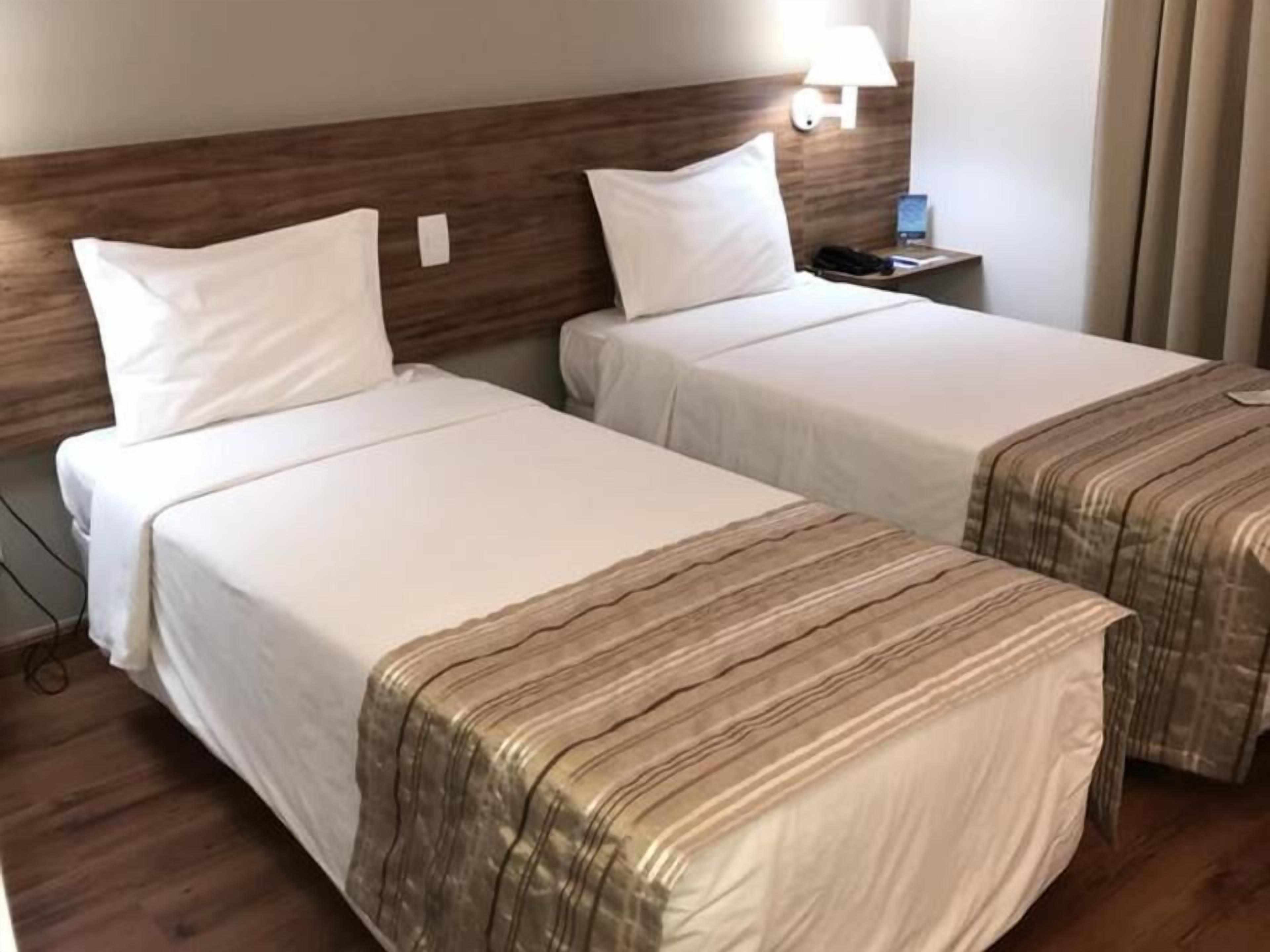 Deux lits simples avec du linge de lit blanc, une tête de lit en bois, une table de chevet avec une lampe et un téléphone.