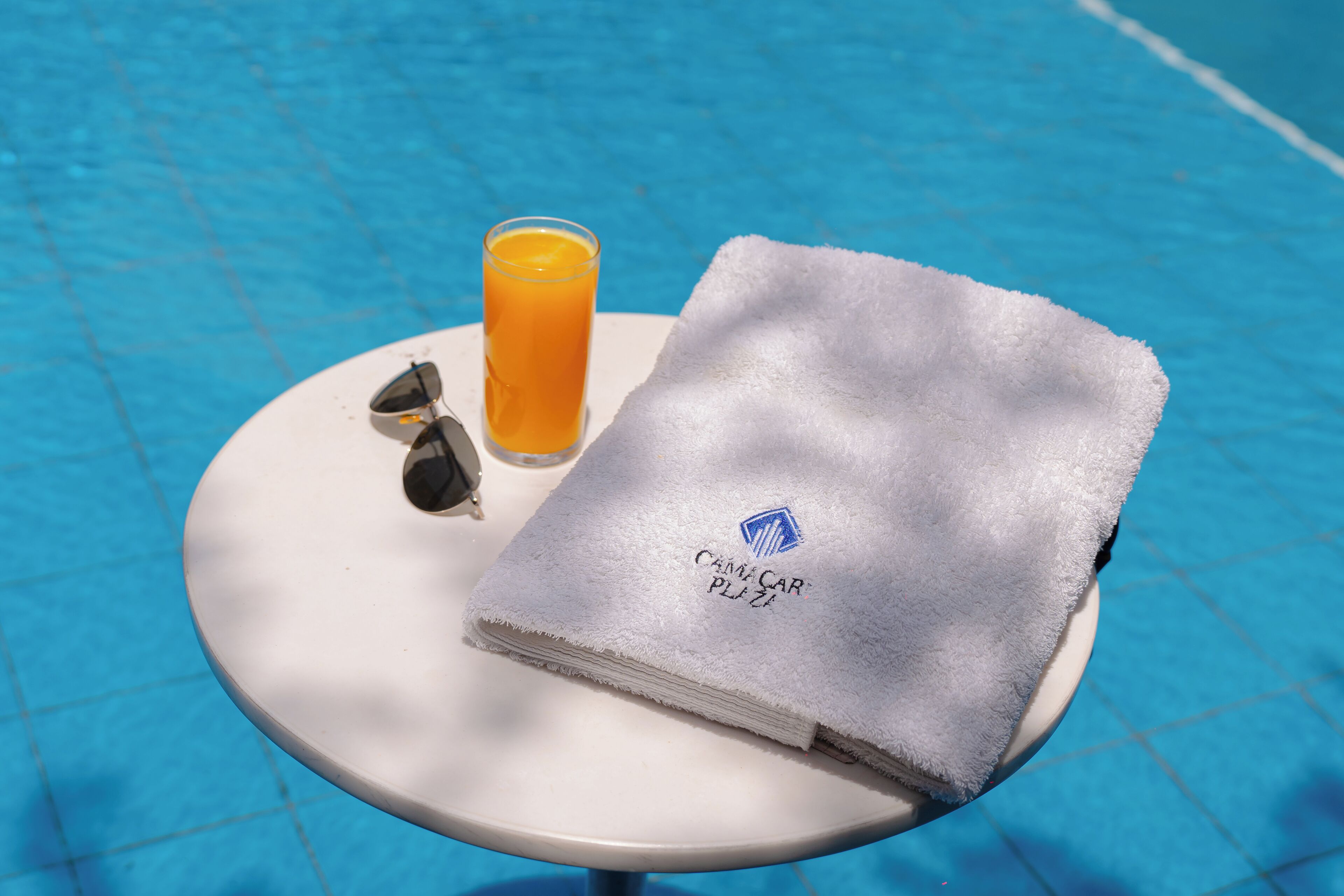 A white table with a folded towel and sunglasses, and a glass of orange juice by a pool.
