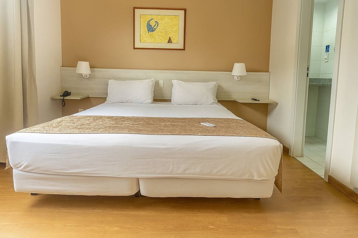 Una habitación de hotel con una cama grande, una mesita de noche, una lámpara de pared y una obra de arte enmarcada sobre la cama.