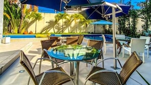 Un área junto a la piscina con una mesa de vidrio y sillas de mimbre, rodeada de sombrillas y palmeras.