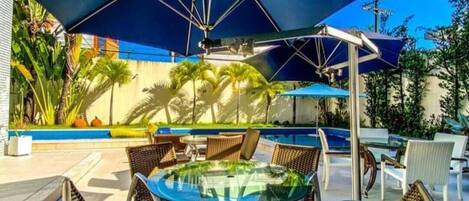 Un espace aménagé au bord de la piscine, avec une table en verre et des fauteuils en osier, entouré de parasols et de palmiers.