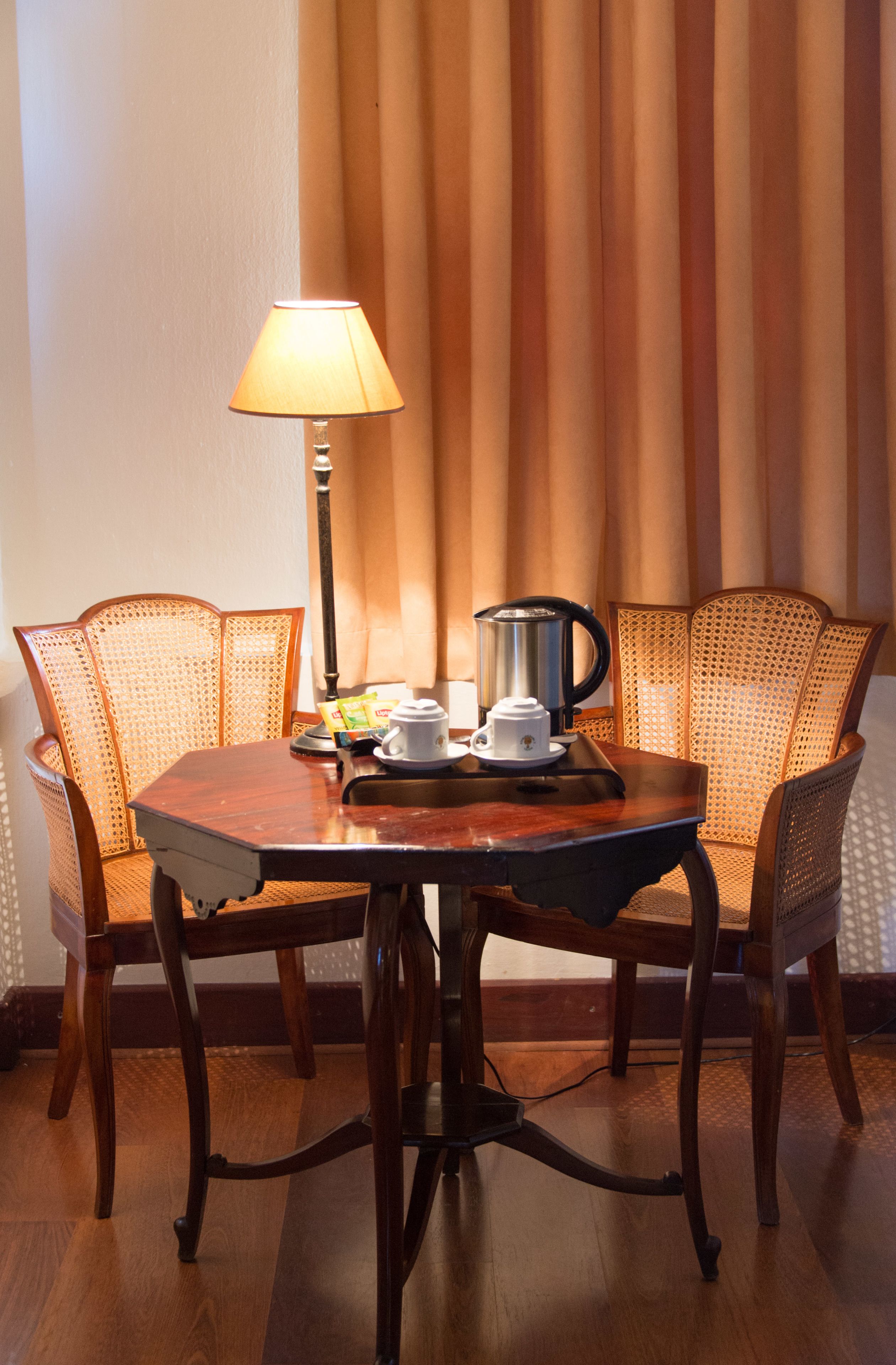 Una mesa de madera para comer con una lámpara, una cafetera y una taza. Hay sillas de mimbre alrededor de la mesa.
