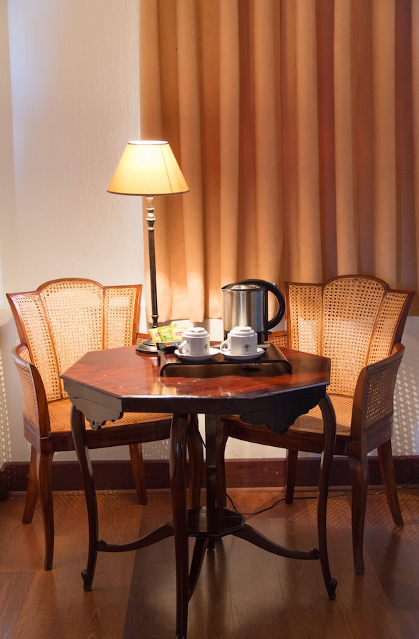 Una mesa de madera para comer con una lámpara, una cafetera y una taza. Hay sillas de mimbre alrededor de la mesa.