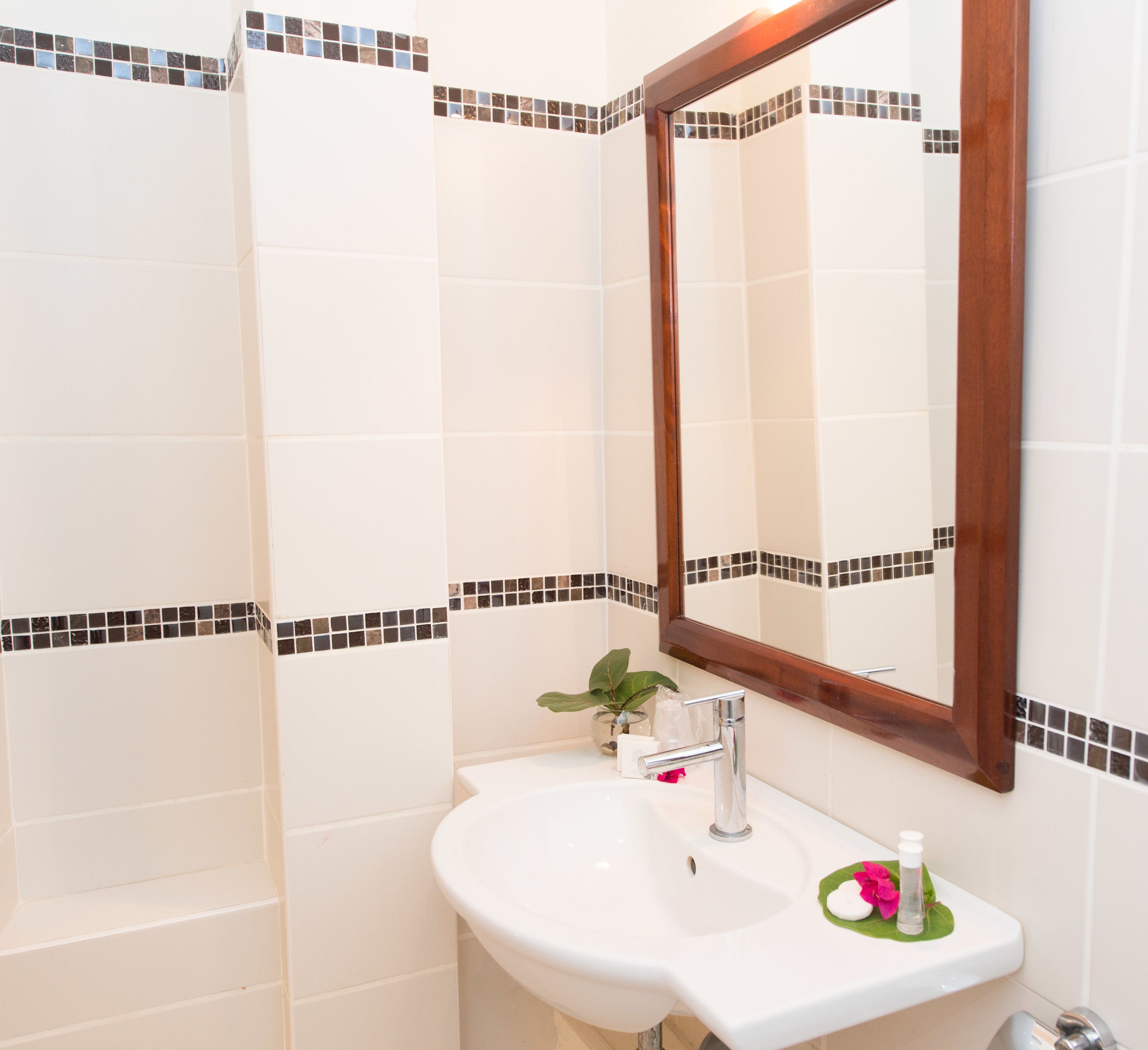 A bathroom with white tiles, a wooden-framed mirror, a sink, and a potted plant.