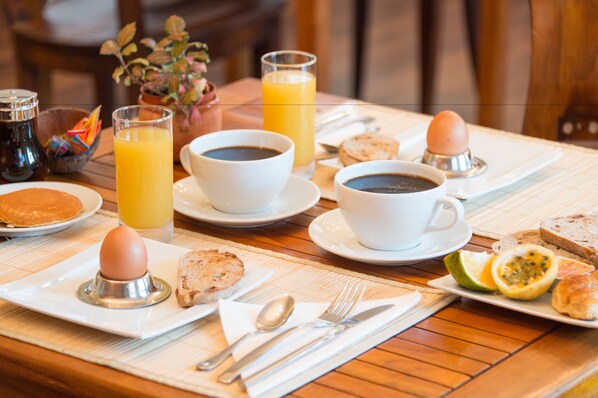 Una mesa de desayuno con platos de huevos, tostadas y fruta, junto con tazas de café y jugo de naranja.