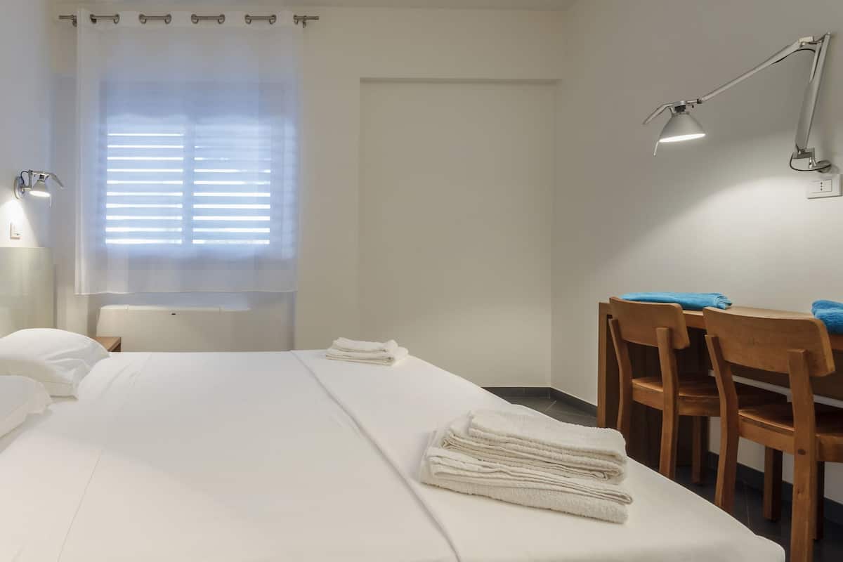 Une chambre d’hôtel avec une tête de lit en bois, un lit avec du linge de maison blanc, un bureau avec des chaises et une lampe fixée au mur.