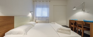 Quarto de hotel com cabeceira de madeira, cama com lençóis brancos, escrivaninha com cadeiras e uma luminária fixada na parede.