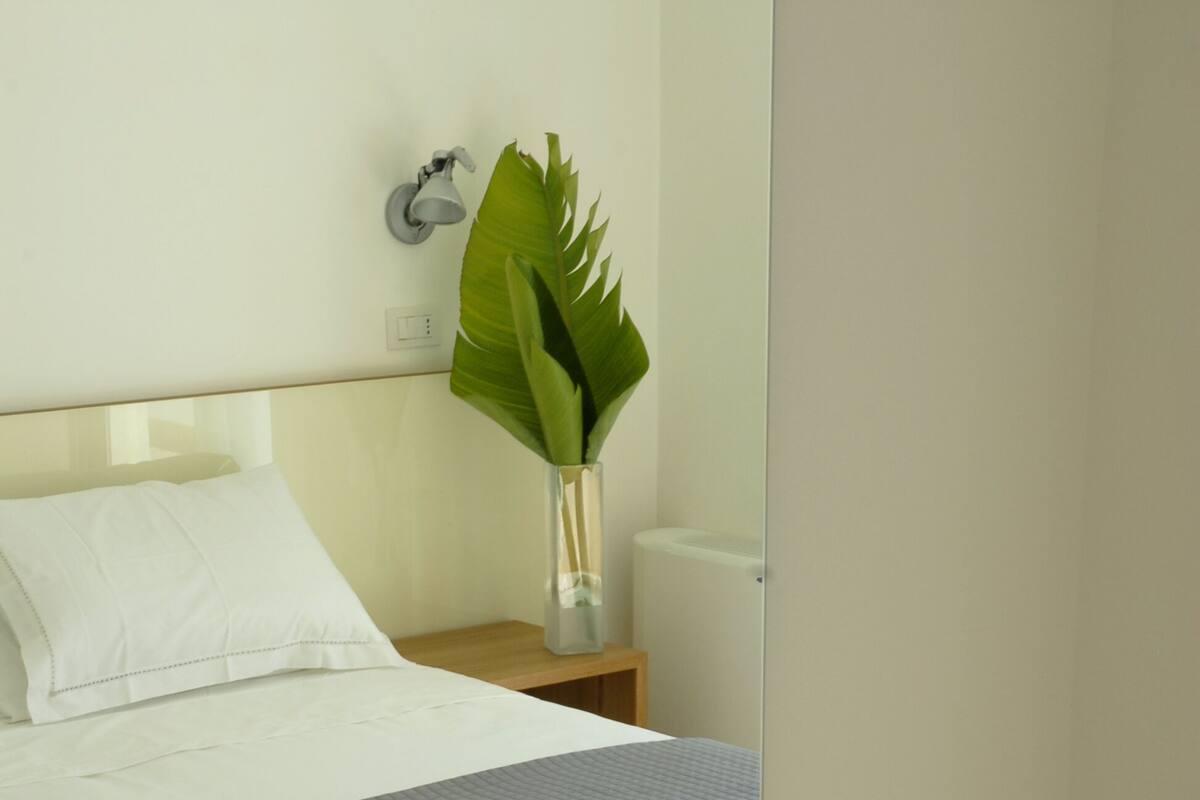 Un lit bien fait, avec du linge de lit blanc et une couverture grise, une table de chevet en bois avec une lampe et une plante, et une chaise en bois dans une pièce au style moderne.