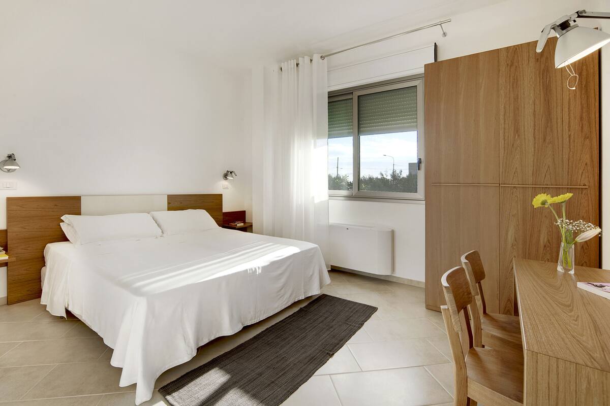 Une chambre d’hôtel moderne dotée d’un grand lit, de tables de chevet, d’une armoire en bois, d’un bureau avec une chaise et d’une fenêtre offrant une vue agréable.