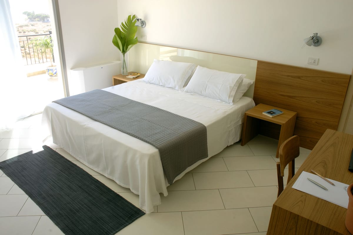 Une chambre d’hôtel bien aménagée, avec un grand lit, une table de chevet avec une lampe, un petit bureau avec une chaise et une vue sur l’extérieur par une fenêtre.