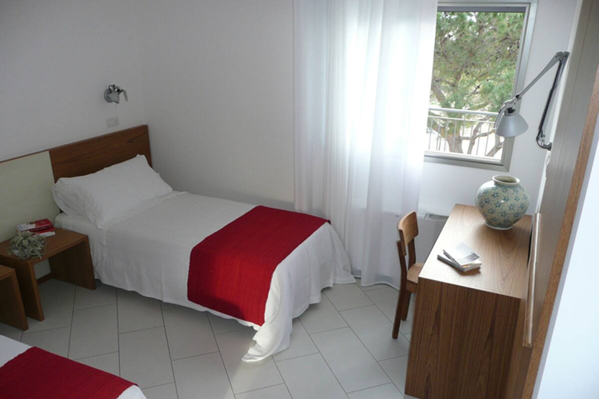 Une chambre à coucher comprenant un lit, un bureau, une chaise, une table de chevet et une fenêtre donnant sur des arbres.