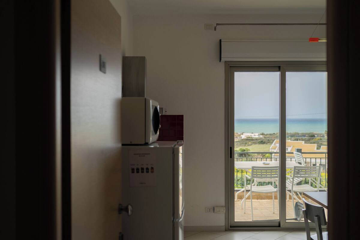 Une cuisine équipée d’un réfrigérateur et d’un four à micro-ondes, ainsi qu’une porte-fenêtre coulissante donnant sur un balcon avec vue sur la mer.