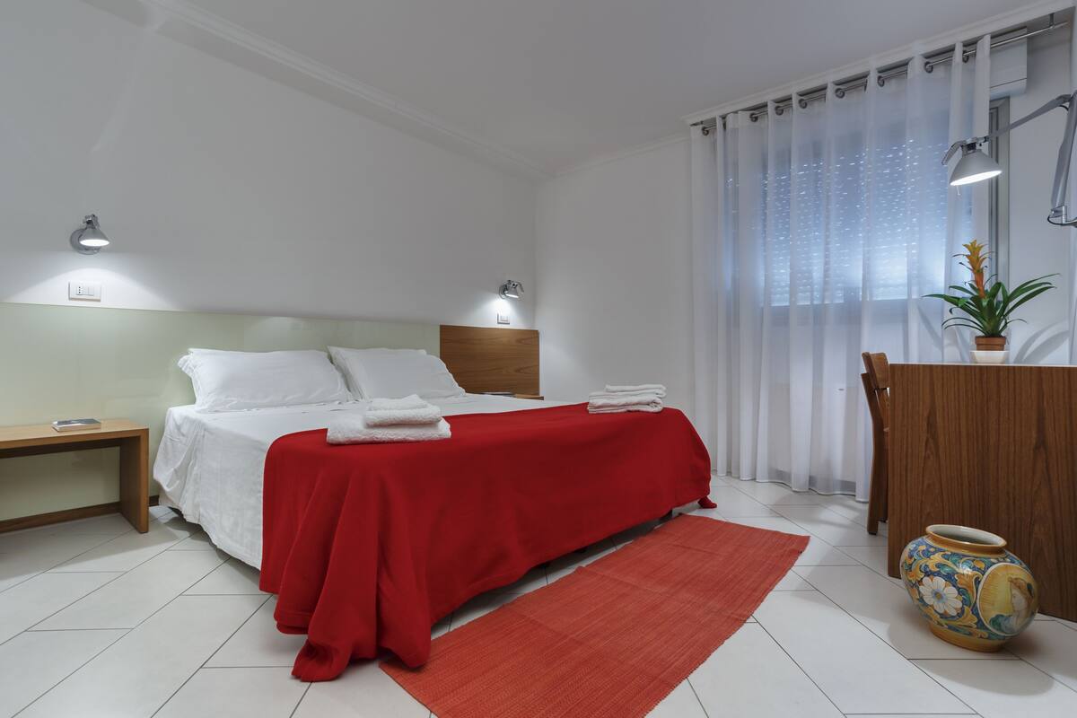 Une chambre à coucher bien rangée, avec un lit, des tables de chevet, une commode, un tapis rouge, une commode en bois et une plante en pot.