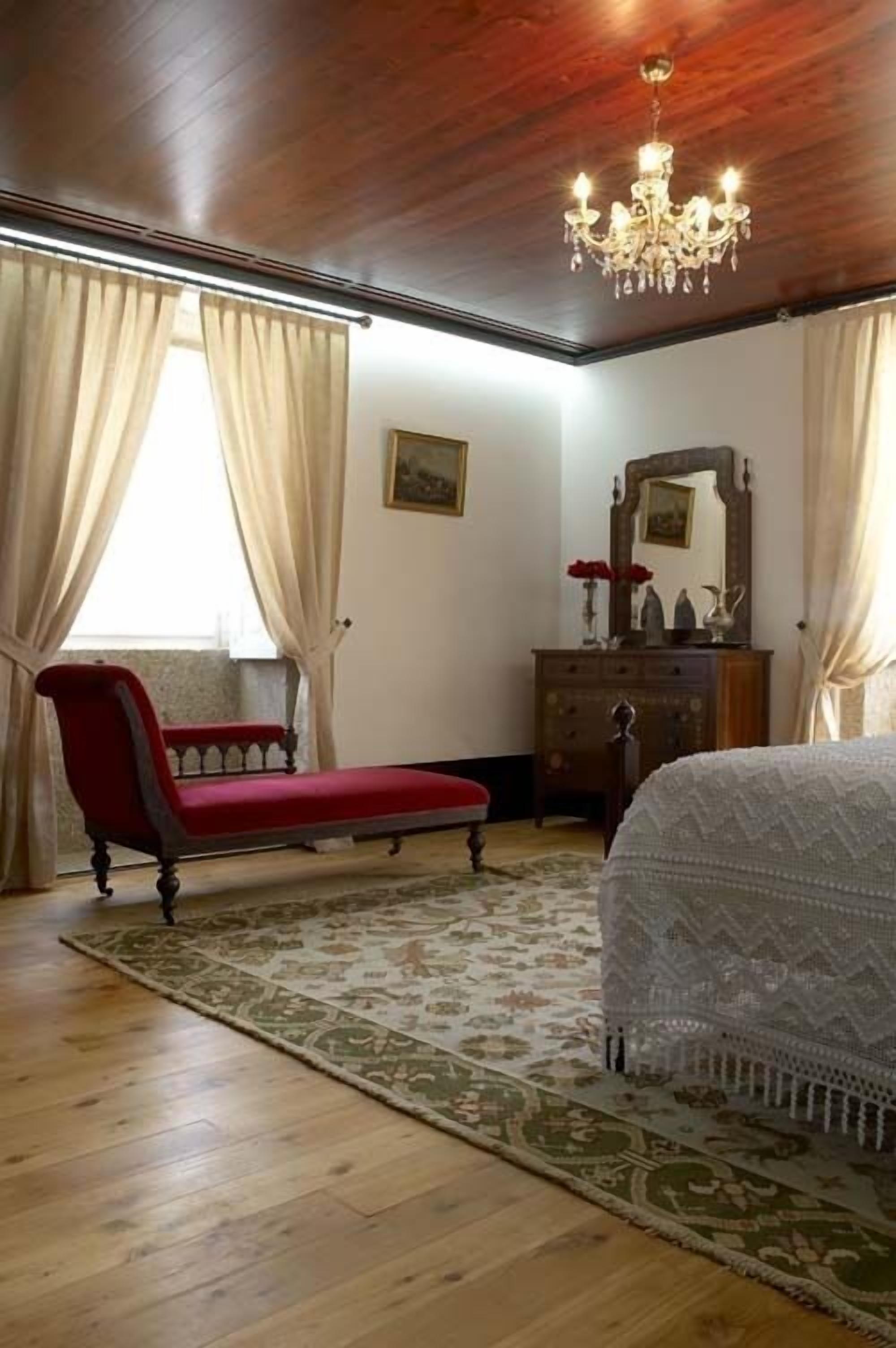 A bedroom with a wooden ceiling, a chandelier, a red sofa, a white bed, a wooden dresser, and a patterned rug.