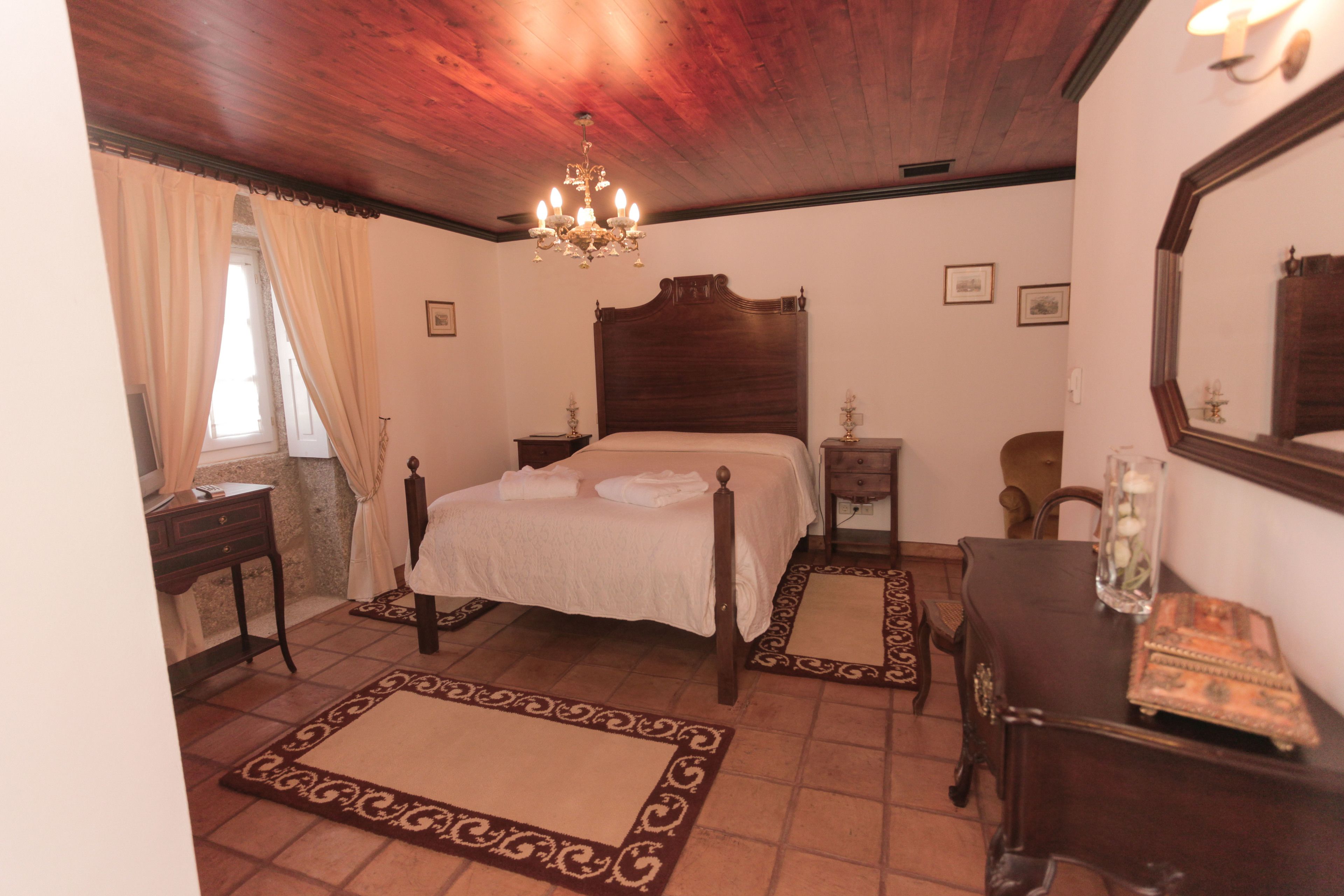 A bedroom with a wooden ceiling, a large bed with a headboard, a chandelier, a dresser, and a mirror.