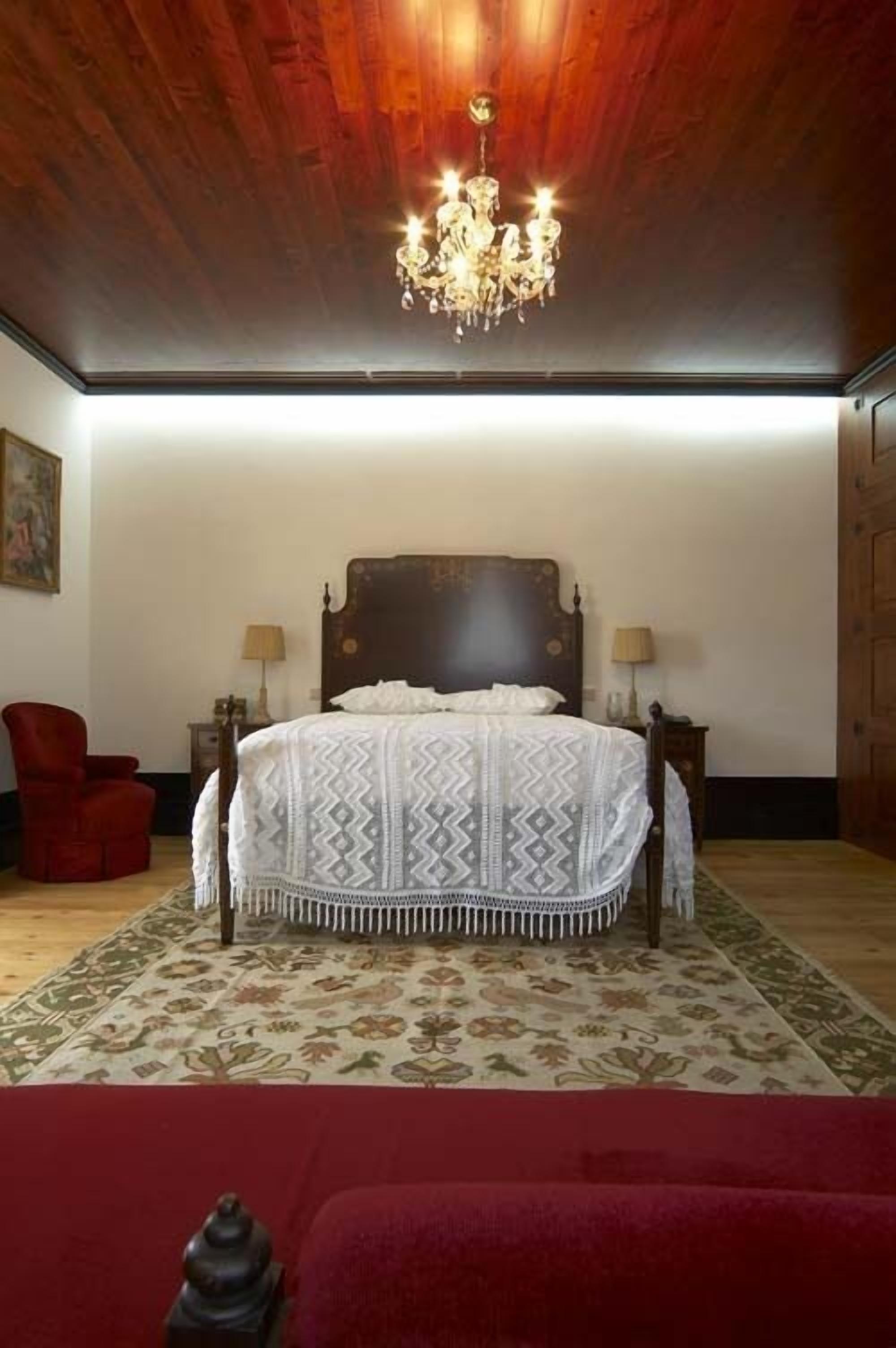A bedroom with a wooden ceiling, a chandelier, a patterned bedspread, a red chair, and a red sofa.