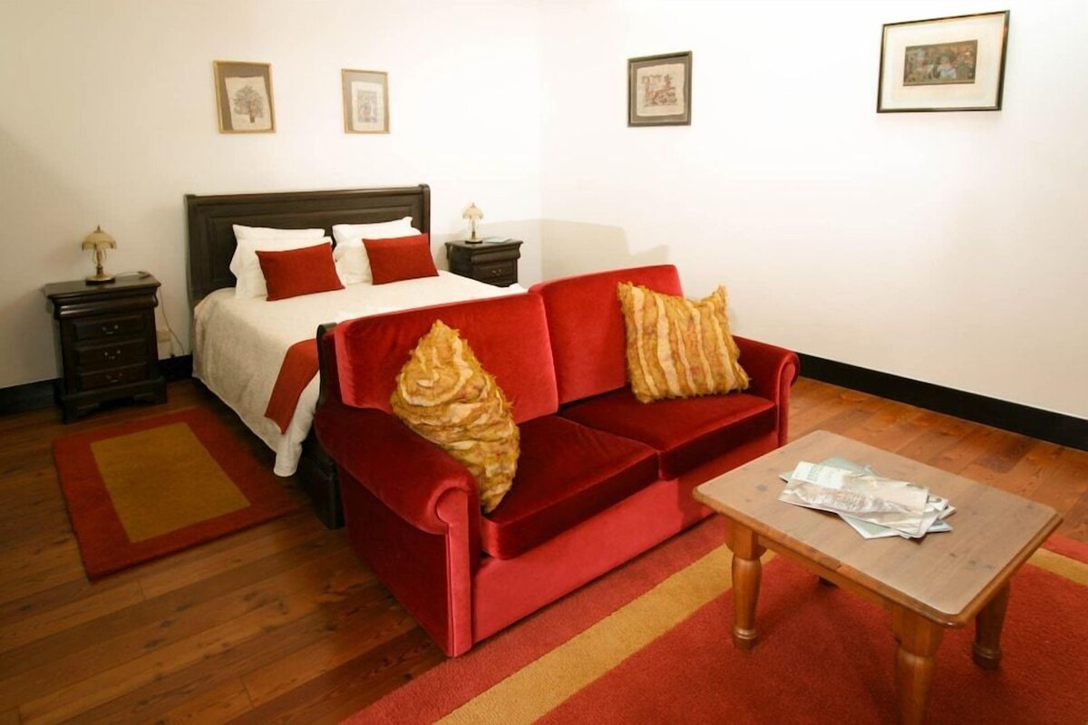 A bedroom with a bed, a red sofa, a wooden table, and framed pictures on the wall.