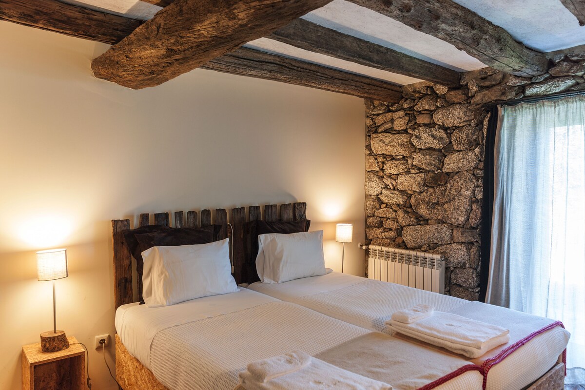 Uma cama de casal com lençóis brancos, cabeceira de madeira, parede de pedra e mesa de cabeceira com abajur.