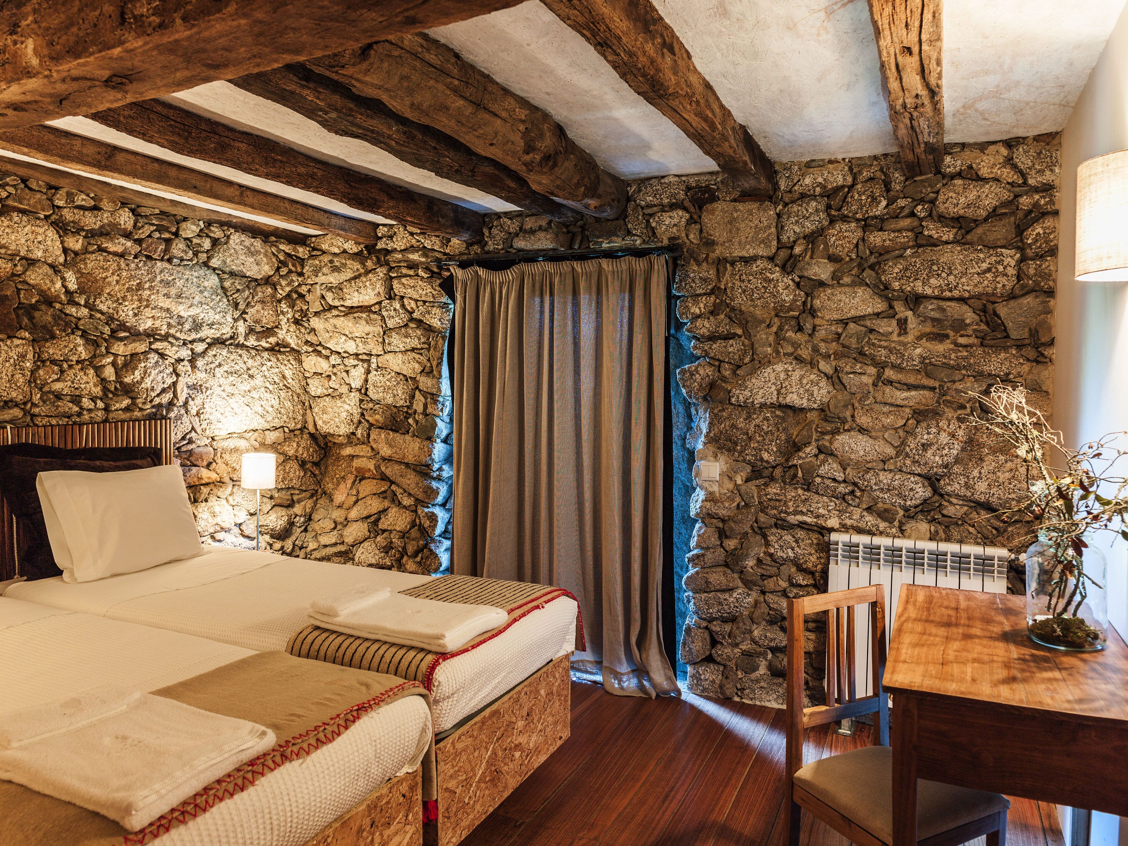 A bedroom with a stone wall, wooden beams, a bed with white bedding, a wooden table with a lamp, and a chair.