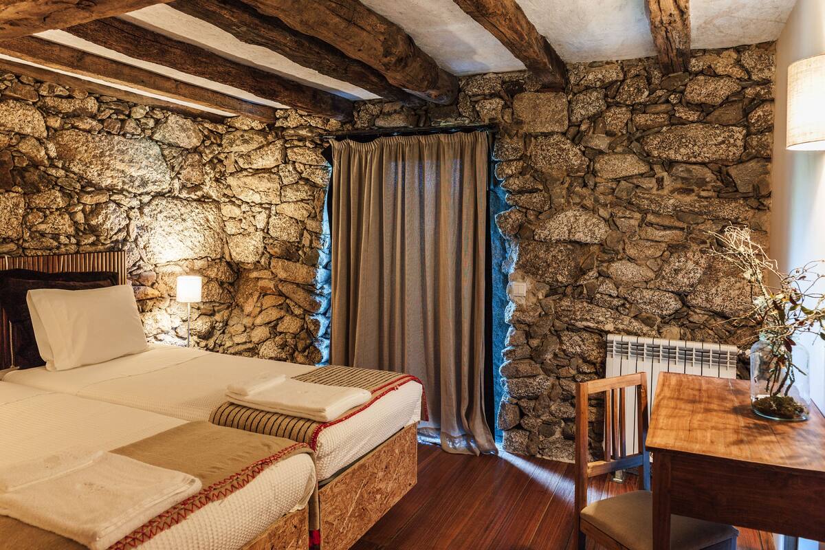 Een slaapkamer met een stenen muur, houten balken, een bed, een stoel, een tafel en een lamp.