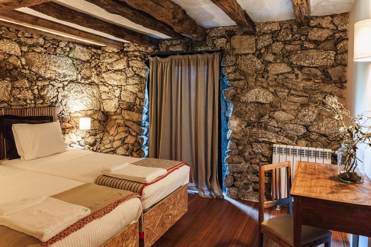 Een slaapkamer met een stenen muur, houten balken, een bed, een stoel, een tafel en een lamp.