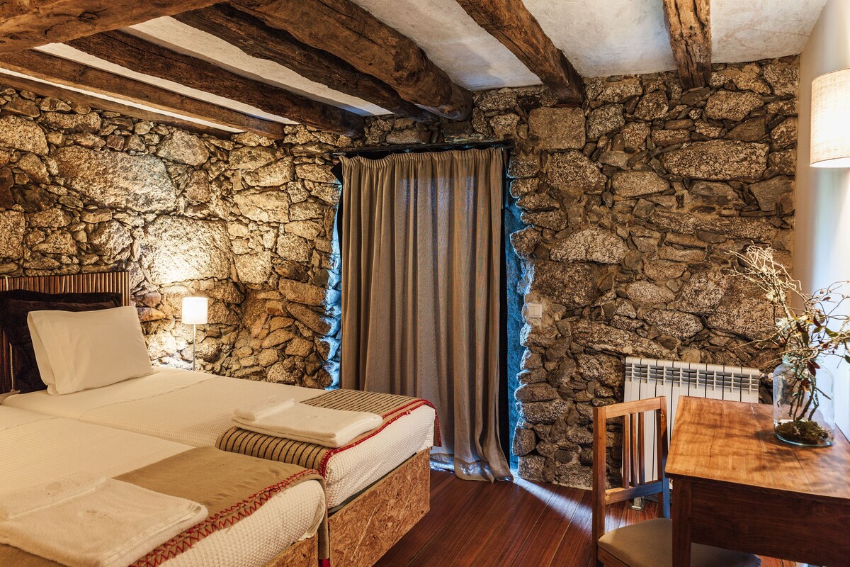 Quarto com parede de pedra, vigas de madeira, cama, cadeira, mesa e abajur.