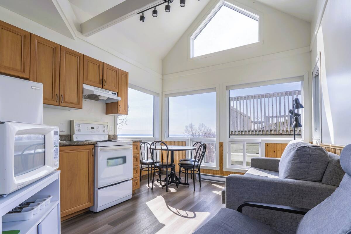 A compact kitchen with wooden cabinets, a microwave, and a stove. There is a dining area with a table and chairs, and a gray sofa in the living area. Large windows provide natural light and views of the outside.