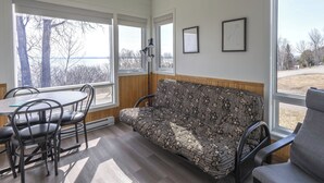 Een gezellige kamer met een houten bank, een tafel met stoelen en grote ramen met uitzicht op bomen en een wateroppervlak.