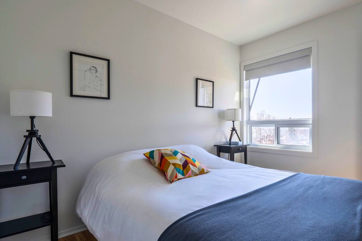 A neatly arranged bedroom with a bed, two bedside tables, a window with blinds, and two framed pictures on the wall.