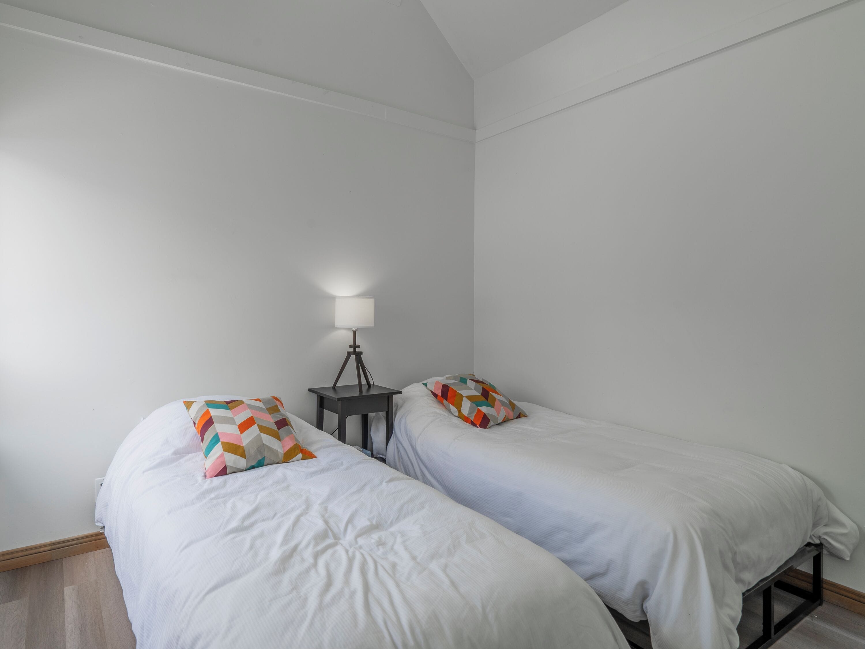 A room with two single beds, each with a colorful geometric pillow, a small bedside table with a lamp, and plain white walls.