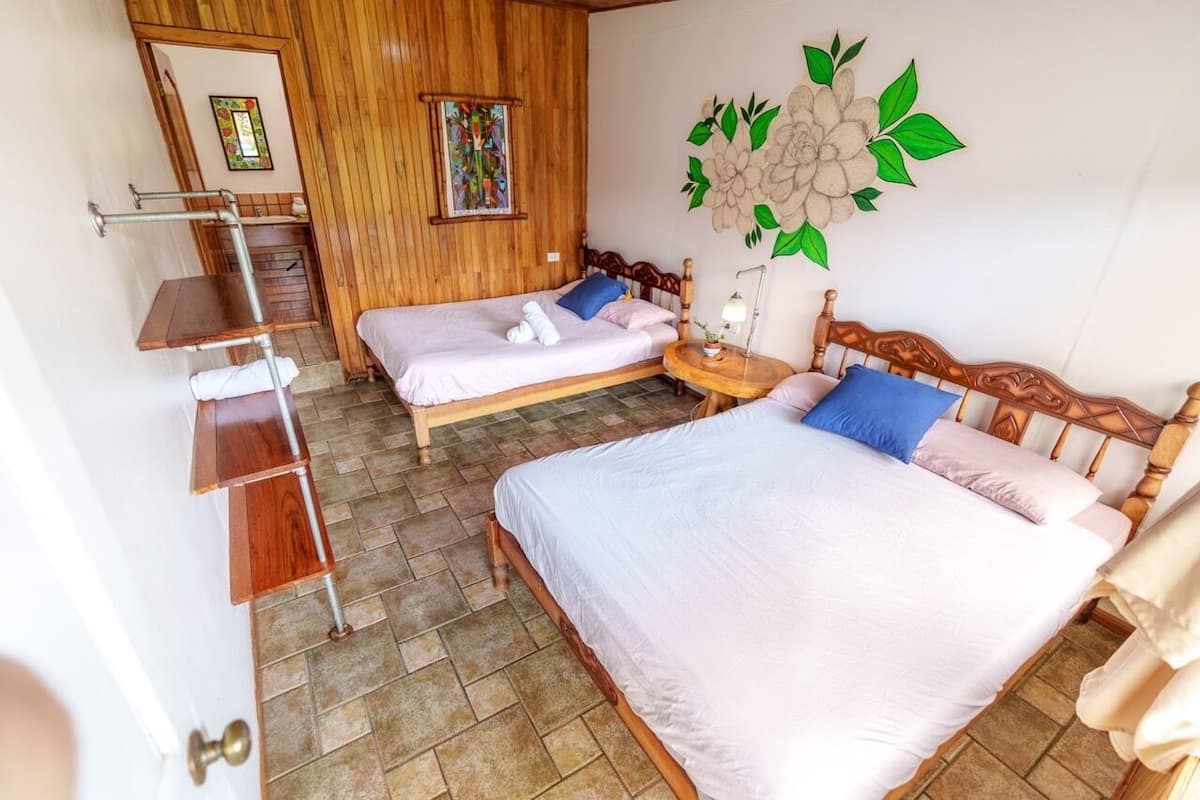 A room with two beds, a wooden wall, a small table with a lamp, and a wall-mounted shelf.