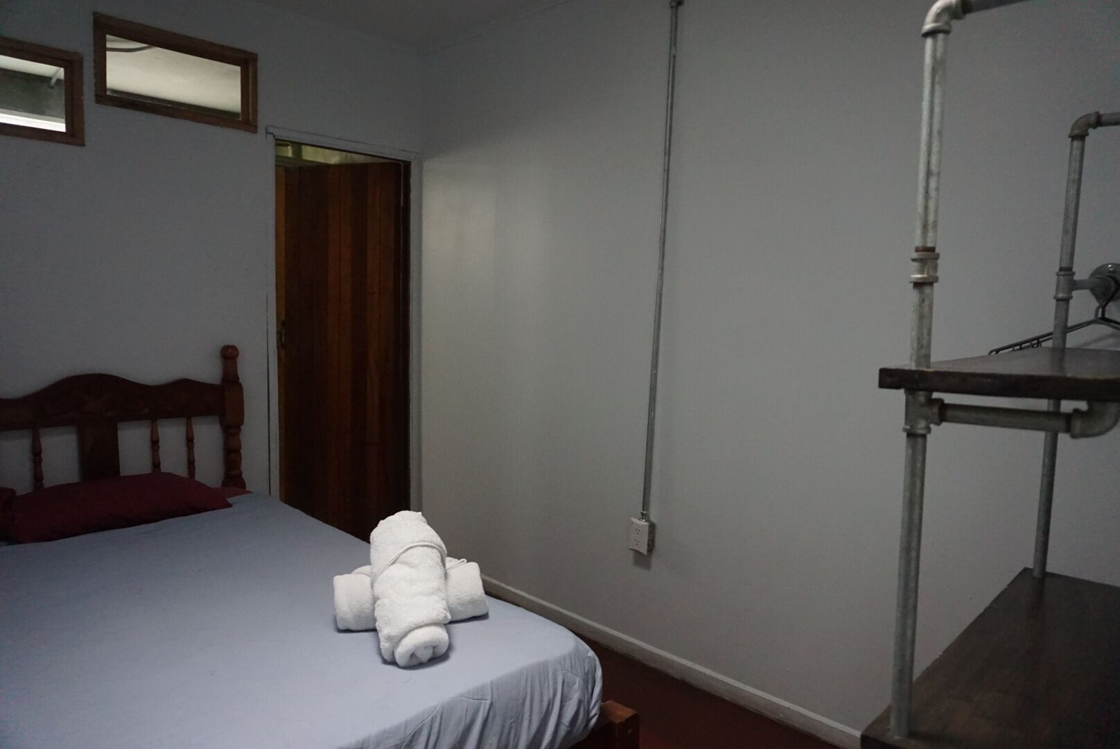 A bedroom with a bed, a wooden headboard, a red pillow, a white towel, a wooden door, and a metal stand with a white object on it.