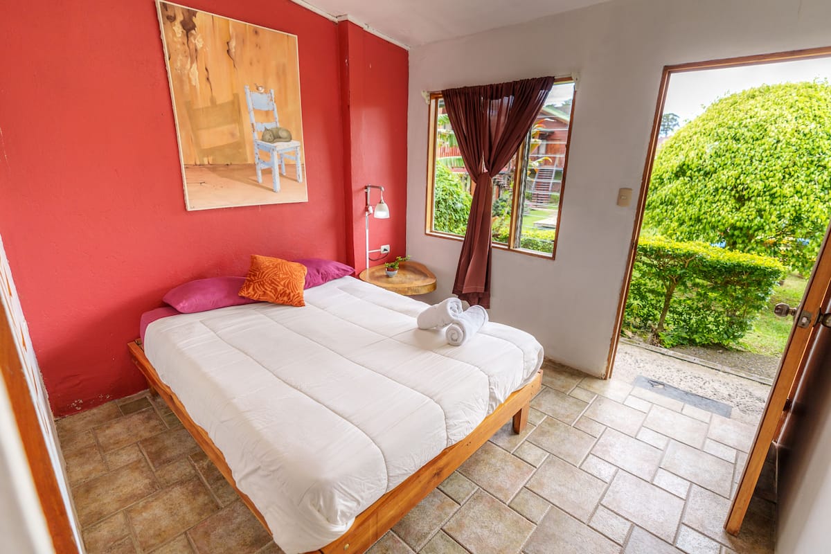 A bedroom with a bed, pillows, a nightstand, a lamp, a window with curtains, and a view of greenery outside.