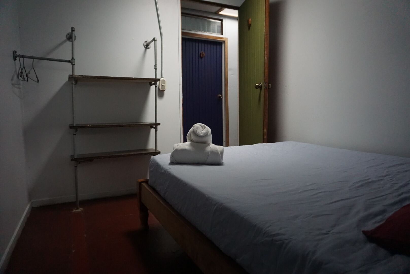 A small, dimly lit room with a single bed, a towel on the bed, a metal rack, and a door with a window.