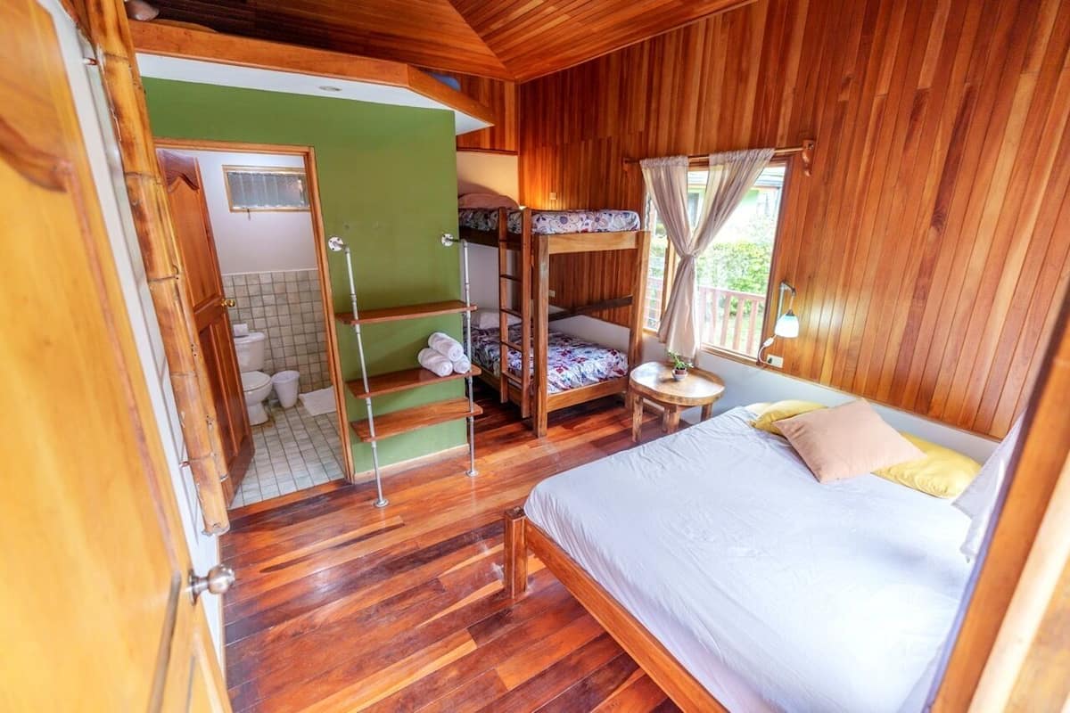 A room with a bunk bed, a single bed, a small round table, and a bathroom with a toilet and sink.