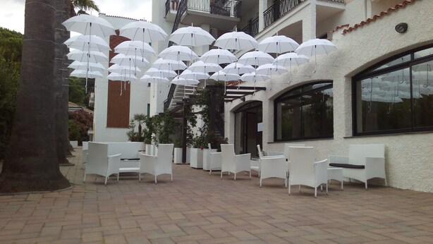 A courtyard with white chairs and umbrellas, a building with a balcony, and a large window. - Residence Riviera (Pietra Ligure)