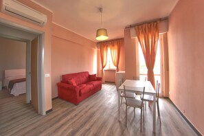 Una habitación con un sofá rojo, una mesa de comedor con sillas, una lámpara en el techo y una ventana con cortinas.