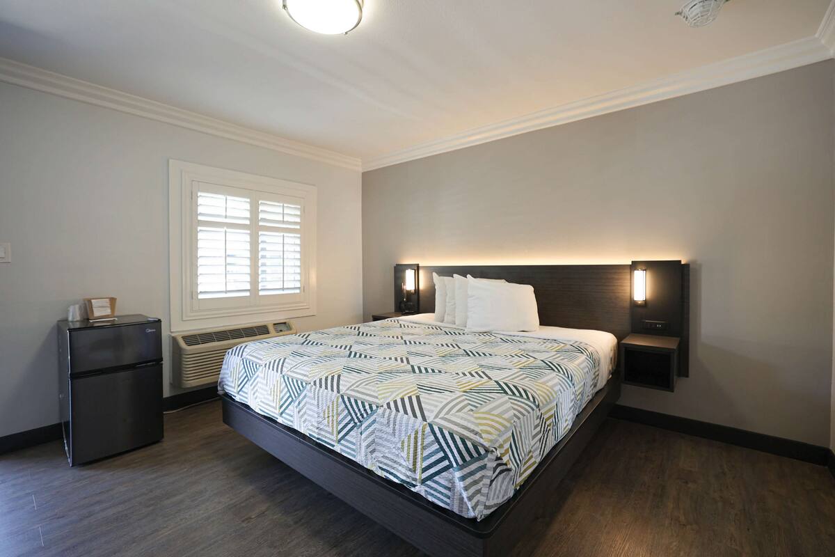 A bedroom with a bed, a small refrigerator, a window with blinds, and wall-mounted lights.